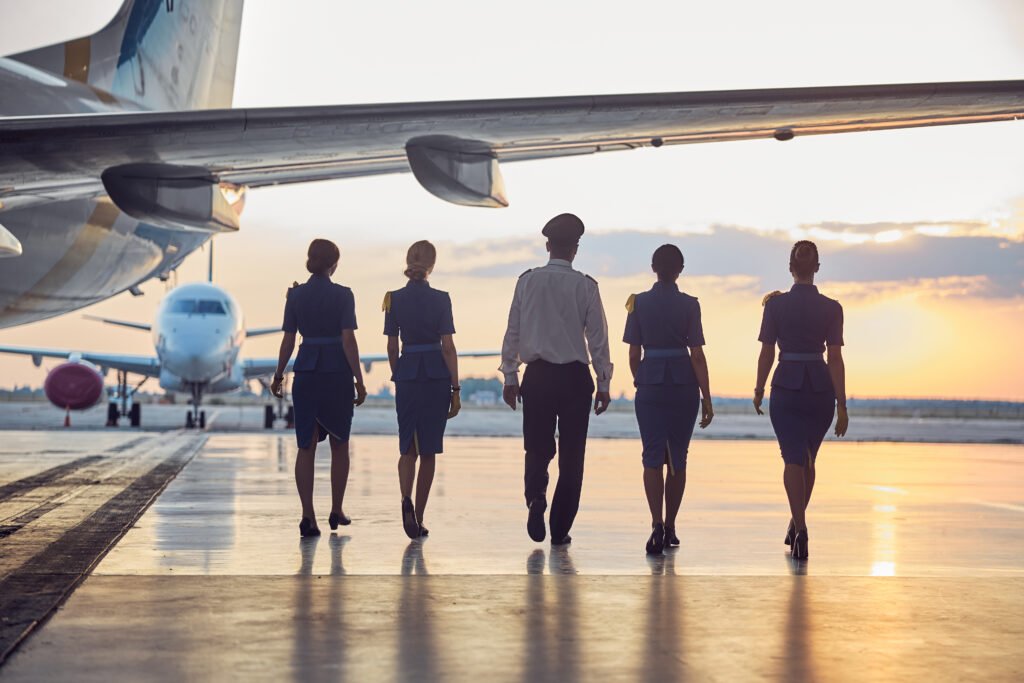 Full length portrait of four stewardess in business uniform with handsome pilot walking through the airport near the aircraft. Aviation Recruitment demand.