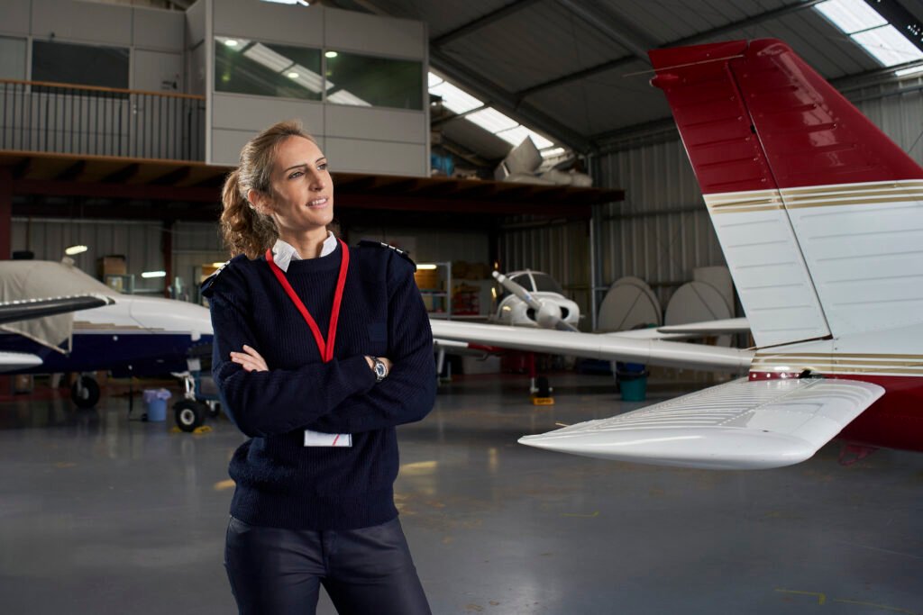 Young pilot posing smiling in the hangar surrounded by airplanes. Women in Aviation Recruitment